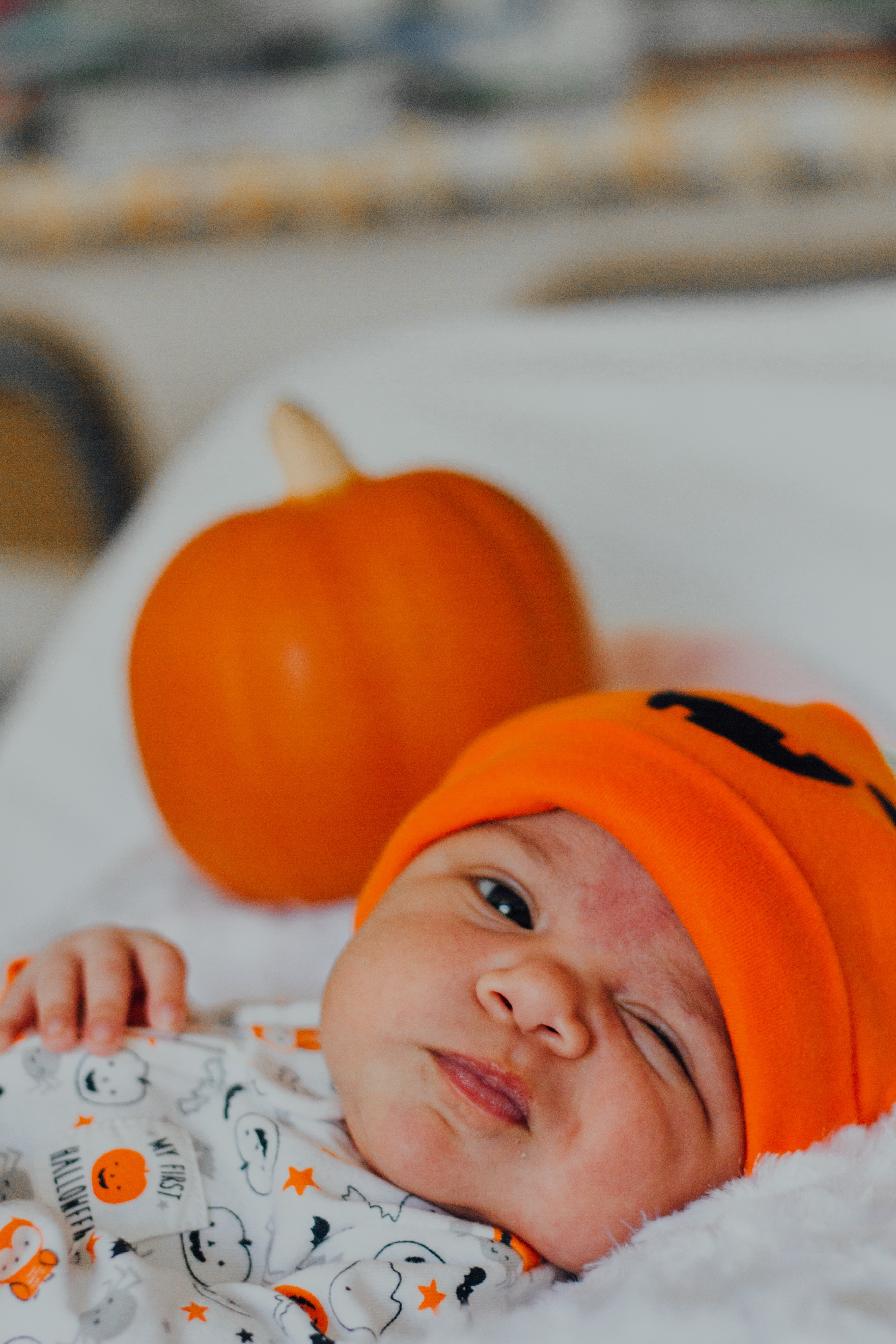 Baby dressed in a Halloween onesie and hat with a pumpkin behind him,  