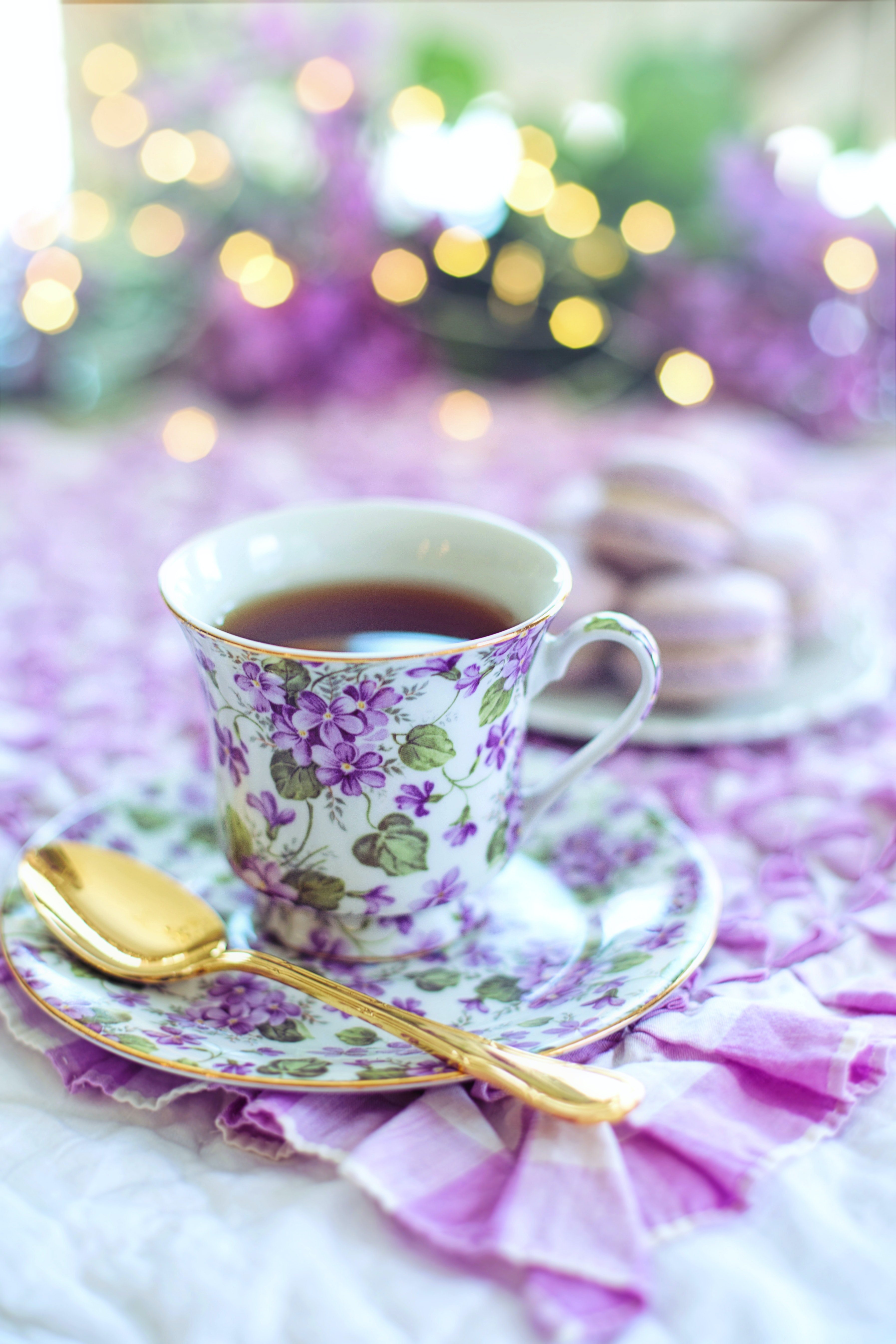 Ceramic or Porcelain Tea cup and plate with purple flowers on it and a golden spoon resting against the plate.  In the slightly blurred background is a plate of light pink macarons and sparkly lights.  
