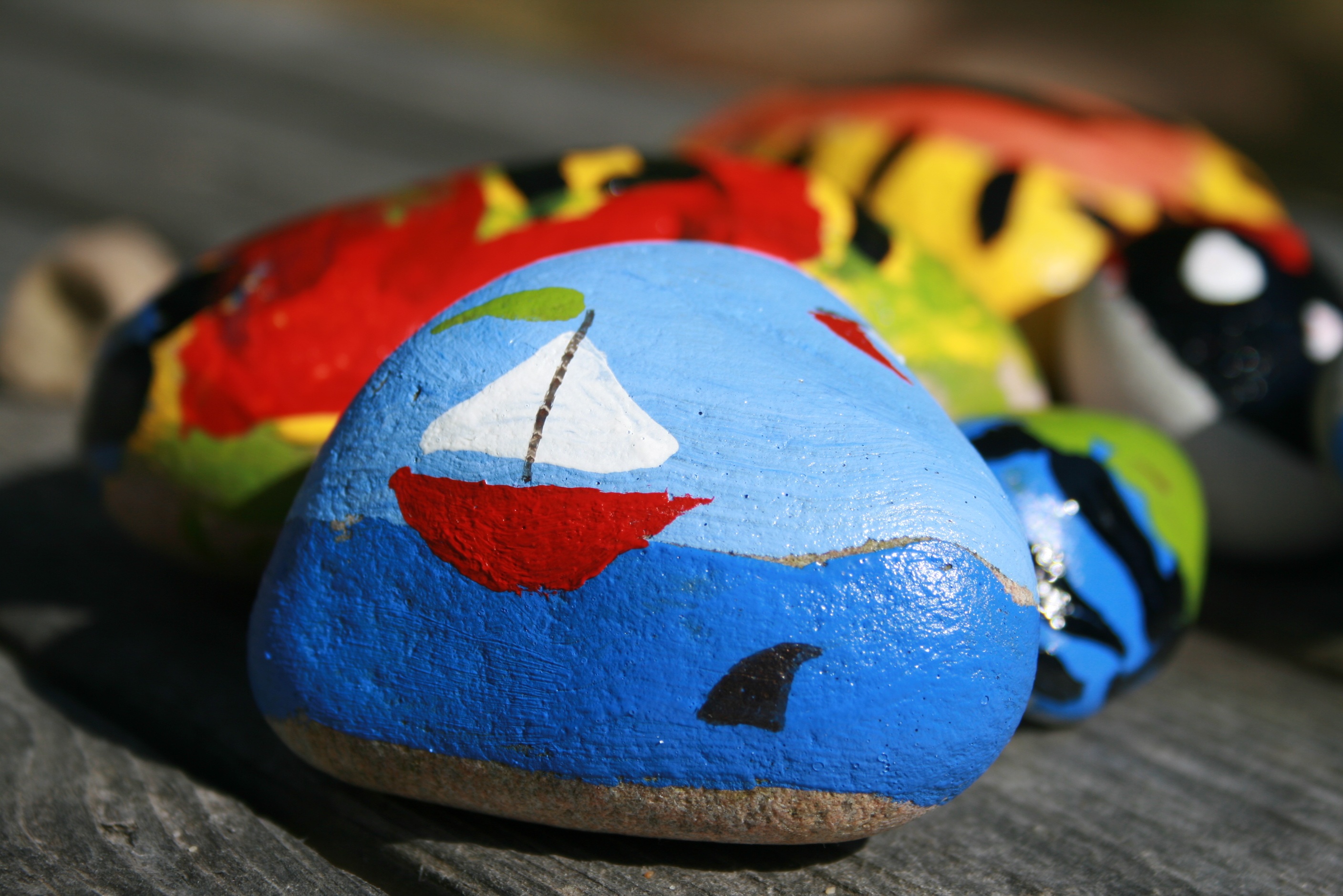 Rock painted to look like a boat on the ocean with a shark fin sticking out of the water.  Other painted rocks are behind it. 