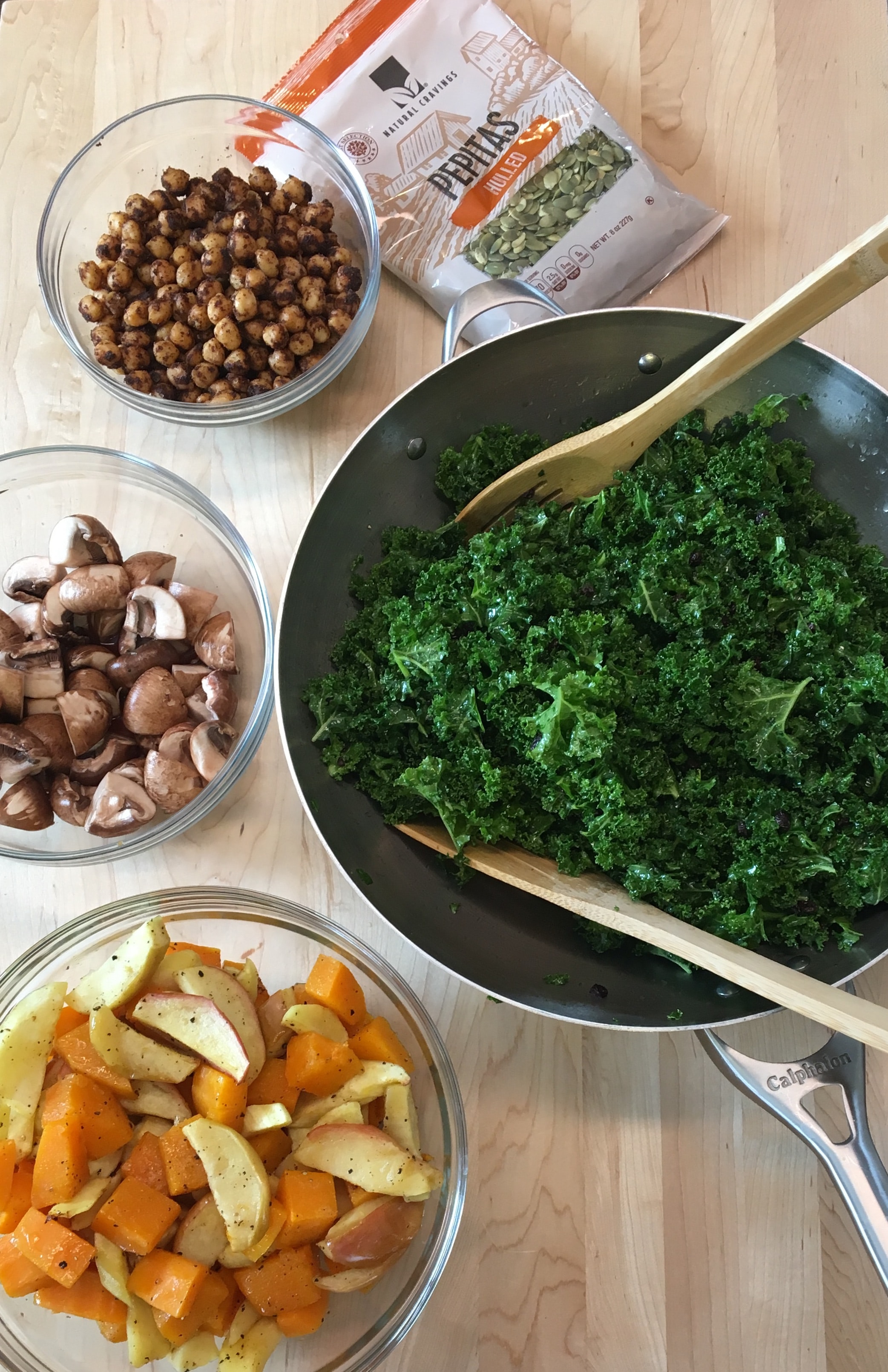 Photo of ingredients for a Kale Salad including  a bowl of kale, a bag of pepitas, a bowl of seasoned chickpeas, a bowl of raw mushrooms, and a bowl of mixed cooked apples and butternut squash.