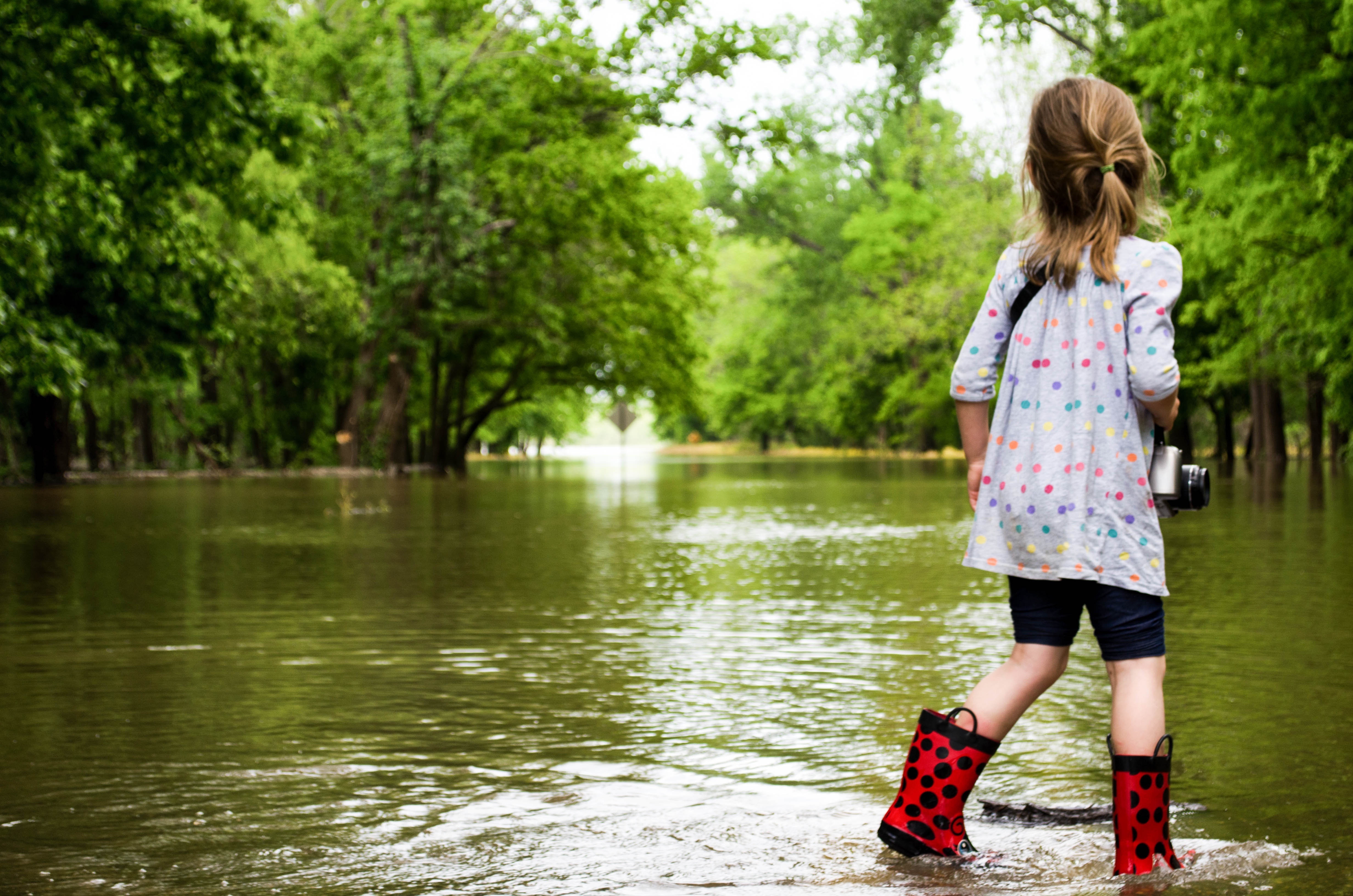 Photo of a young girl in boots standing in shallow water surrounded by trees.