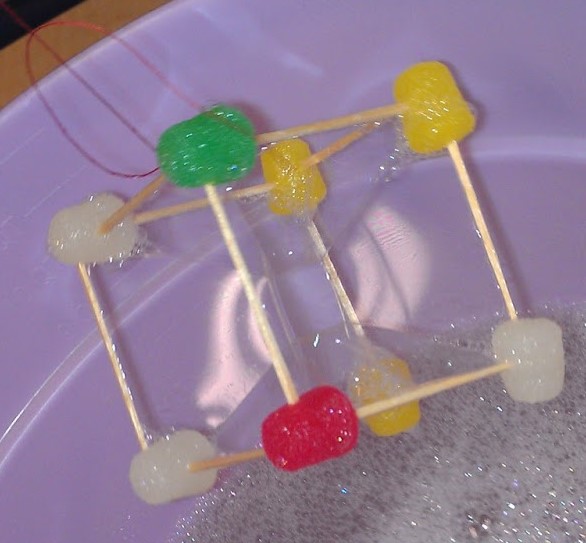 Gumdrops attached to each other with toothpicks in the shape of a cube being pulled out of a soapy solution in a purple bowl.