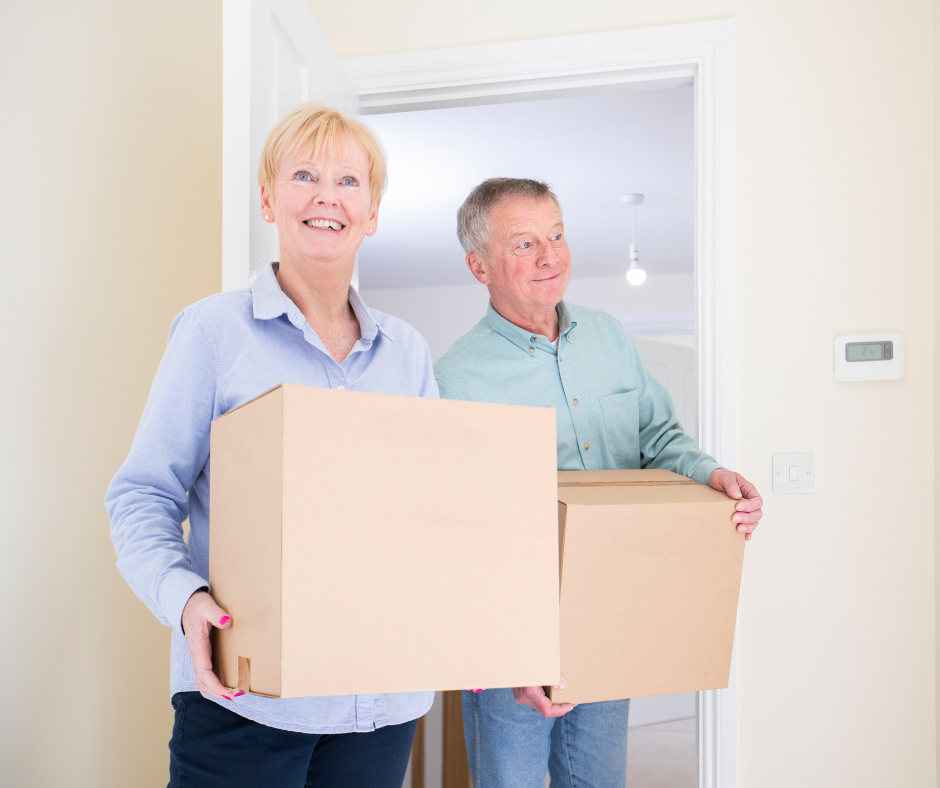 An older woman and man holding boxes walking through a door into a white room.