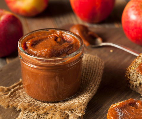 Small jar of apple butter surrounded by apples and apple butter on a spoon and bread/