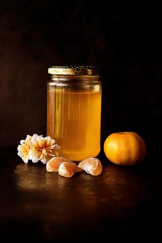 A jar of honey, flowers, orange, and orange segments with a dark brown background.