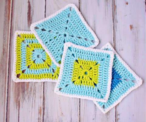 Four overlapping crochet squares in shades of lime green, white, and blue on a white washed wood background.