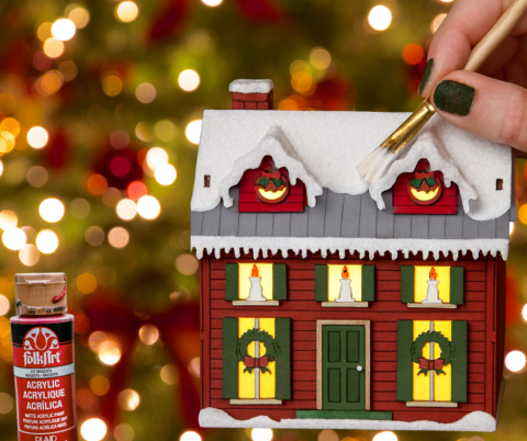 Hand painting a wooden house over Christmas tree background and a bottle of paint.