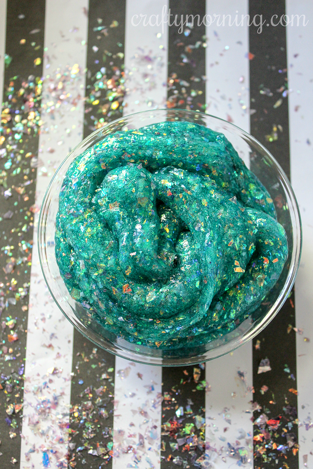 Teal sparkly slime in a glass bowl on a black and white striped background.