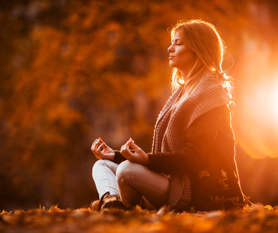 Woman in Fall clothing sitting on the ground meditating with the sun in the background.