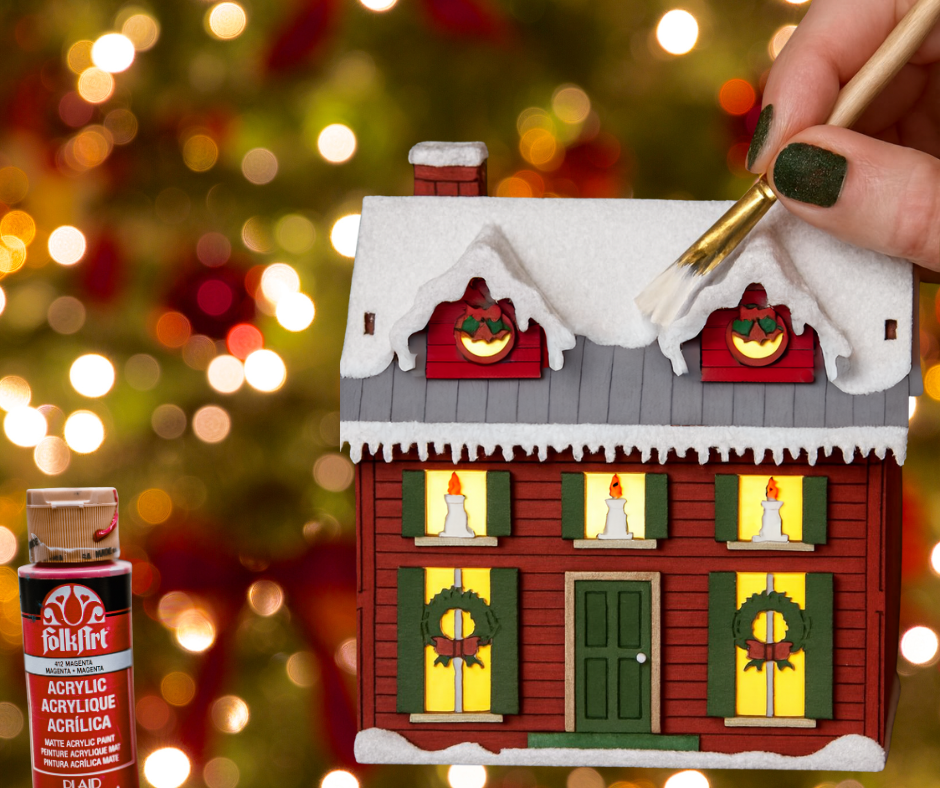 Hand painting a wooden house over Christmas tree background and a bottle of paint.